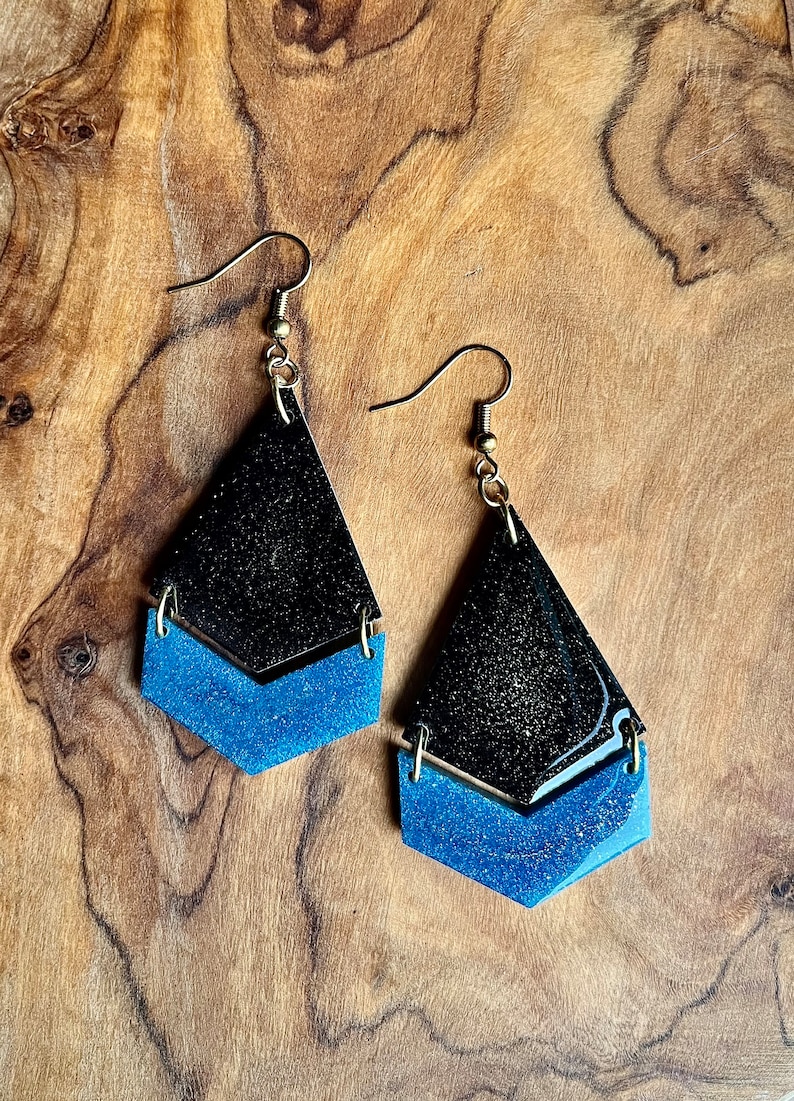 K&ouml;nnte beinhalten: Ein Paar Ohrh&auml;nger mit schwarzem und blauem Glitzer-Finish. Die Ohrringe sind tropfenf&ouml;rmig mit einer kleineren Tropfenform darunter.