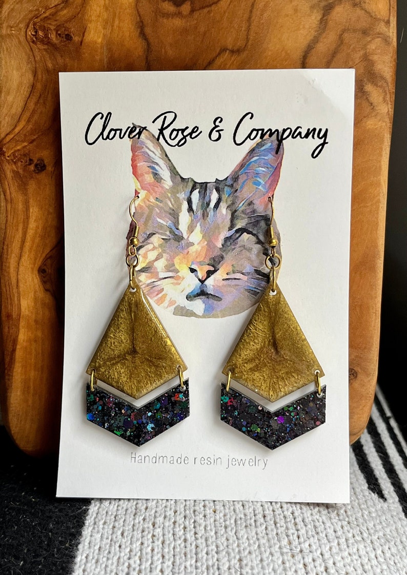 K&ouml;nnte beinhalten: Goldene und schwarze Harzohrringe mit Chevron-Muster. Die Ohrringe sind handgefertigt und haben eine Glitzer-Oberfl&auml;che. Die Ohrringe werden auf einer Karte mit einer Katzenillustration und dem Text "Clover Rose & Company" und "Handgefertigte Harzschmuck" pr&auml;sentiert.