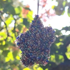 Op de afbeelding: Een stervormig ornament gemaakt van kleurrijke kralen, voornamelijk blauw, groen en rood. Een donker touwtje is bevestigd om op te hangen. Ideaal voor feestdecoratie of als uniek cadeau.