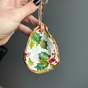 May include: A gold-painted oyster shell ornament with a holly and berry design. The ornament is decorated with green holly leaves and red berries on a white background.