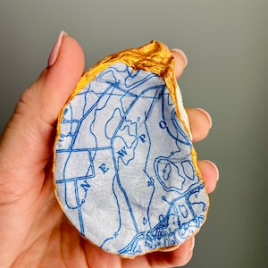May include: A white oyster shell with a gold rim, decorated with a blue and white map print. The map shows a body of water with the letters "W.P." and "E."