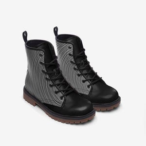 May include: A pair of ankle-high lace-up boots featuring a solid black toe cap, heel, and eyelet panel. The side panels display a grey and black wavy line pattern. The boots have black laces, black pull tabs, and a thick brown lugged rubber sole.