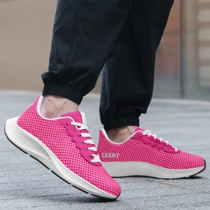 May include: A pair of bright pink sneakers featuring a small checkered pattern, white laces, and a white sole. The word EXERT is printed in white on the side of the shoe. The sneakers are worn with black pants, showcasing a low-top athletic design.