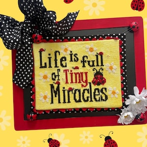 Puede incluir: Un bordado enmarcado con la frase "Life is full of Tiny Miracles" sobre un fondo amarillo. El marco es rojo y negro, adornado con detalles de mariquitas y un lazo de lunares blancos y negros. El fondo es amarillo con diseños de margaritas y mariquitas.