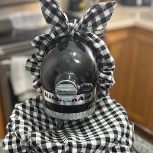 May include: A black KitchenAid stand mixer with a black and white checkered fabric cover and bow. The cover drapes over the mixer and has a ruffled edge. The bow is on top of the mixer.