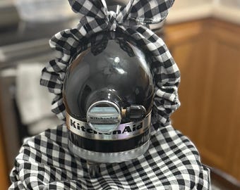 May include: A black KitchenAid stand mixer with a black and white checkered fabric cover and bow. The cover drapes over the mixer and has a ruffled edge. The bow is on top of the mixer.