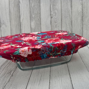 Op de afbeelding: Een herbruikbare voedselafdekking met een bloemenpatroon. De afdekking is gemaakt van stof en heeft een dieprode achtergrond met roze, witte en turquoise bloemen en bladeren. Het is ontworpen om over een rechthoekige glazen schaal te passen.
