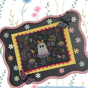 May include: A cross-stitch sampler with a ghost design, framed in a pink and black border. The sampler features a ghost, letters, and floral elements. The text "Ghost Sampler" is at the top, and "Design By: Christina Groneck" is at the bottom.
