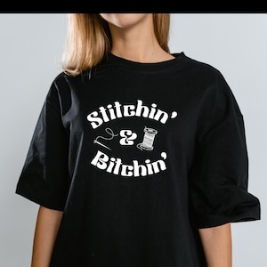 Puede incluir: Camiseta negra con el texto blanco "Stitchin' & Bitchin'" y un gráfico de aguja, hilo y carrete. La camiseta tiene mangas cortas y un corte holgado.
