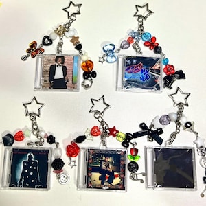 May include: Five miniature CD case keychains with star-shaped clasps. Each keychain features a unique album cover insert and is decorated with a variety of colourful plastic beads, including butterflies, bows, hearts, and musical note charms.