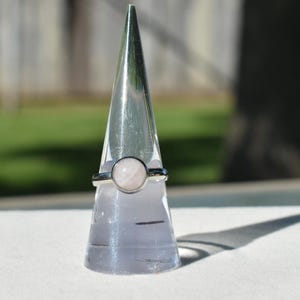 Puede incluir: Un anillo de plata con una piedra preciosa redonda de color rosa pálido. El anillo se muestra sobre un soporte transparente en forma de cono. El fondo está borroso, mostrando hierba verde y un árbol.