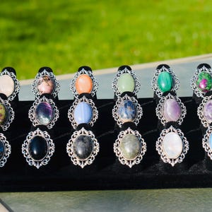 May include: A collection of gemstone rings displayed on a black velvet stand. Each ring features a different oval-shaped stone in various colors, set in an ornate silver-colored metal frame. The background is a blurred green.
