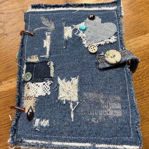 May include: A rectangular journal with a blue denim fabric cover featuring frayed edges, a heart-shaped denim patch, various fabric scraps, lace trim, assorted buttons, and a metal ring binding. A small fabric strap with a floral button secures the cover.