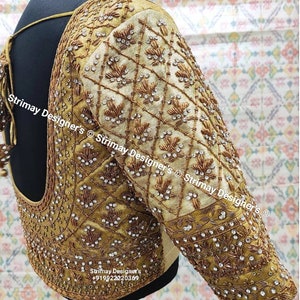 Puede incluir: Una blusa bordada en oro y blanco con un escote en V profundo y mangas largas. La blusa está decorada con intrincados abalorios y lentejuelas. El texto "Strimay Designer's" es visible en la blusa.