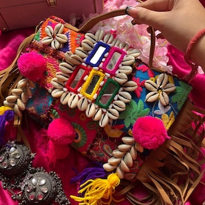 May include: A colorful patchwork handbag with a fringe trim, adorned with seashells, mirrors, and pom-poms. The bag has a brown leather strap and is made of a variety of fabrics.