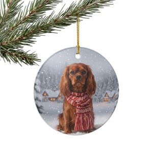 May include: A round ceramic Christmas ornament featuring a Cavalier King Charles Spaniel wearing a red and white scarf. The dog is set against a snowy winter scene with houses and trees. The ornament hangs from a gold string.