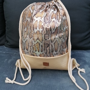 Könnte beinhalten: Eine beige Kordeltasche mit Schlangenhautmuster. Die Tasche hat einen braunen Lederboden und eine weiße Kordel.