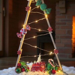 Könnte beinhalten: Eine Weihnachtsbaumdekoration aus Holz mit Zuckerstangen- und Schleifenornamenten, roten Sternakzenten und einem Miniatur-Roten Zug. Der Baum wird von einer Lichterkette beleuchtet und steht auf einer weißen Kunstfelloberfläche.