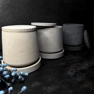 Könnte beinhalten: Drei zylindrische Beton-Pflanzgefäße in verschiedenen Grautönen. Jeder Topf hat einen passenden Deckel und steht auf einem runden Sockel. Die Töpfe sind auf einer dunklen Oberfläche angeordnet, mit einem Zweig blauer Beeren im Vordergrund.