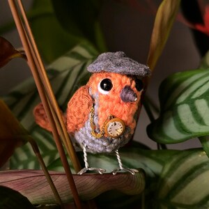 May include: A crocheted bird figurine with orange and grey yarn, wearing a small grey felt cap. The bird features black bead eyes, a dark grey beak, and thin silver wire legs. A gold-toned chain with a miniature round clock is attached.