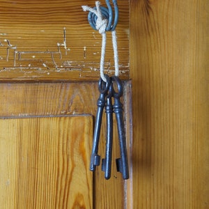 Könnte beinhalten: Drei antike Schlüssel, die an einer Schnur hängen, die an einer Holztür befestigt ist. Die Schlüssel sind aus Metall und haben eine dunkle Patina.