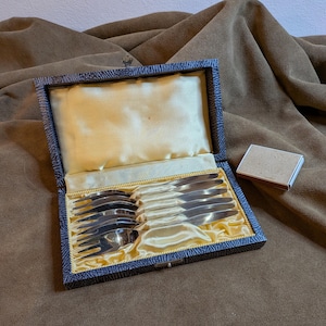 Könnte beinhalten: Ein offenes, antikes Besteckset in einer blau-gold gemusterten Schachtel. Die Schachtel enthält mehrere silberne Gabeln und Löffel, die auf einem gelben Satinfutter liegen. Rechts befindet sich eine kleine, rechteckige Schachtel.