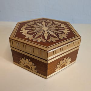 Könnte beinhalten: Eine sechseckige Holzbox mit einem dekorativen Blumendesign auf Deckel und Seiten. Die Box ist aus dunkelbraunem Holz mit helleren Holzakzenten gefertigt. Der Deckel hat ein Sonnenstrahlenmuster.