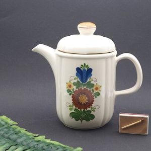 Könnte beinhalten: Weiße Keramik-Teekanne mit einem Blumendesign in den Farben Blau, Braun, Gelb und Grün. Die Teekanne hat einen Deckel und einen Henkel.