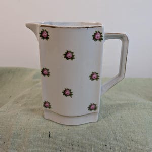 Könnte beinhalten: Weißer Keramikkrug mit rechteckiger Form und Henkel. Der Krug ist mit rosa Rosenblütenmustern und goldenen Verzierungen verziert. Er hat einen Ausguss zum einfachen Ausgießen. Ein dekoratives Vintage-Stück.