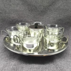 Könnte beinhalten: Ein Set aus sechs silberfarbenen Glasbechern und einer silberfarbenen Zuckerdose mit schwarzem Deckel, alles auf einem silberfarbenen Tablett angeordnet.