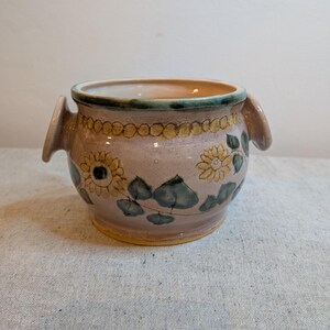 Könnte beinhalten: Ein Keramiktopf mit hellrosa Grundfarbe, verziert mit gelben Sonnenblumen und grünen Blättern. Der Rand ist grün, und der Topf hat zwei kleine Griffe. Der Topf hat einen Durchmesser von etwa 15 cm.