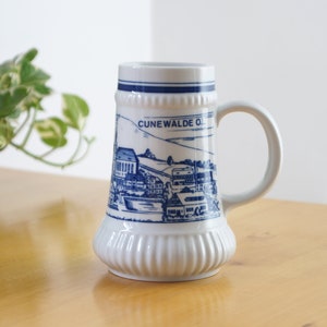 Könnte beinhalten: Weißer Keramik-Bierkrug mit einer blauen und weißen Illustration einer Stadt mit dem Text "CUNEWALDE OL" auf der Seite.
