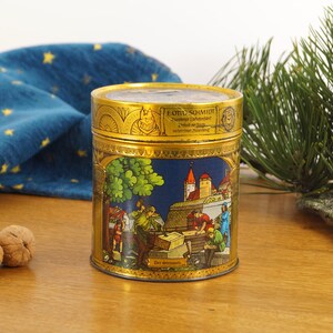 Könnte beinhalten: Eine vintage goldene Dose mit einer blauen und weißen Szene einer mittelalterlichen Stadt mit Menschen, die an einem Gebäude arbeiten. Die Dose trägt den Text "E. Otto Schmidt" und "Nurnberg, Deutschland".