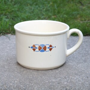 Könnte beinhalten: Weiße Keramiktasse mit einem blauen und braunen geometrischen Muster. Die Tasse hat eine abgerundete Form und einen Henkel.