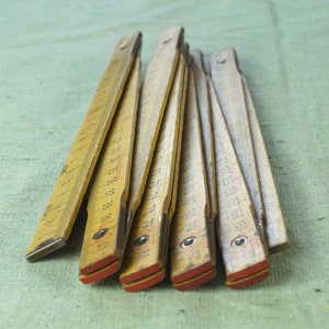 Könnte beinhalten: Ein alter, zusammenklappbarer Holz-Zollstock mit schwarzen und roten Markierungen. Der Zollstock besteht aus hellem Holz und hat rot lackierte Enden. Der Zollstock ist teilweise geöffnet und zeigt die Maße in Zentimetern.