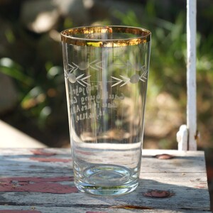 Könnte beinhalten: Ein klares Glas Trinkglas mit goldfarbenem Rand und geätztem Design. Das Glas hat eine weiße Inschrift und ein Muster aus Pfeilen und Kreisen.