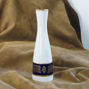 Könnte beinhalten: Eine weiße Keramikvase mit goldfarbenem Rand und einem blauen und goldenen Musterband um die Mitte. Die Vase steht auf einer braunen Stoffoberfläche.