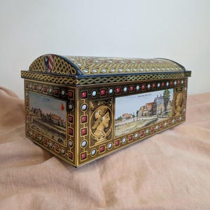 Könnte beinhalten: Eine rechteckige Vintage-Blechdose mit gewölbtem Deckel. Die Dose zeigt detaillierte Kunstwerke, darunter Landschaftsszenen, Porträts und geometrische Muster in Gold, Rot und Braun. Der Deckel hat einen blauen Hintergrund mit goldenen Akzenten.