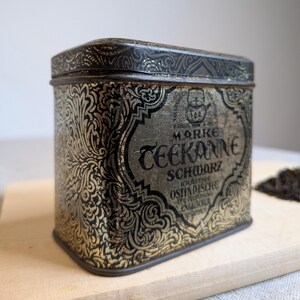 Könnte beinhalten: Eine rechteckige Teedose aus Metall mit einem schwarzen und goldenen floralen Ornamentmuster. Auf der Vorderseite steht der Text MARKE TEEKANNE SCHWARZ KRAFTIGE OSTINDISCHE TEE MISCHUNG ENGLISCHER, mit einem kleinen Teekannen-Symbol.