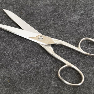 Könnte beinhalten: Offene, silberfarbene Schere mit scharfen Klingen. Die Schere hat ein klassisches Design mit Fingerlöchern und einem Drehpunkt in der Mitte. Die Klingen sind aus glänzendem Metall und die Griffe sind glatt.