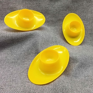 Könnte beinhalten: Drei gelbe Eierbecher aus Kunststoff. Die Eierbecher sind wie eine kleine Schale mit einem breiten Rand geformt.
