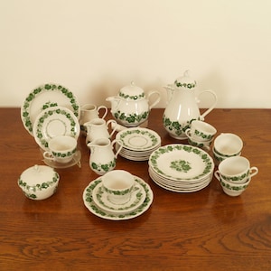 Könnte beinhalten: Ein komplettes weißes Porzellan-Teeservice mit grünem Blättermuster. Das Set umfasst Tassen, Untertassen, Teller, eine Teekanne, einen Sahnekännchen und eine Zuckerdose. Das Teeservice ist auf einer Holzoberfläche angeordnet.