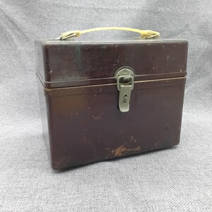 Könnte beinhalten: Eine braune Metallbox mit einem Verschluss und einem gelben Kunststoffgriff. Die Box hat einen abgenutzten, Vintage-Look.