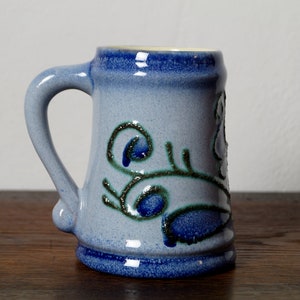 Könnte beinhalten: Eine blaue Keramiktasse mit einem grünen und blauen Blumendesign. Die Tasse hat einen Henkel und einen abgerundeten Boden.