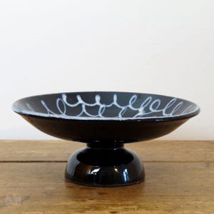 Könnte beinhalten: Eine schwarze Keramik-Fußschale mit einem weißen abstrakten Design. Die Schale hat einen breiten, flachen Boden und einen erhöhten, runden Sockel.