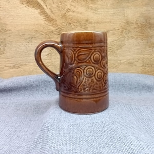 Könnte beinhalten: Eine braune Keramiktasse mit einem erhabenen Blumendesign. Die Tasse hat einen breiten Griff und einen abgerundeten Körper.