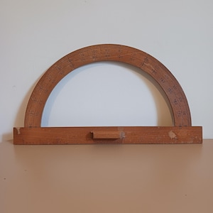 Könnte beinhalten: Ein Vintage-Winkelmesser aus Holz, ein Werkzeug zum Messen von Winkeln, mit einer halbkreisförmigen Skala von 0 bis 180 Grad. Der Winkelmesser aus hellbraunem Holz hat eine gerade Basis mit einem Griff. Die Skala ist in Grad markiert.