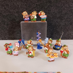 Könnte beinhalten: Eine Sammlung kleiner, farbenfroher Bärenfiguren in verschiedenen Posen. Einige Bären halten Herzen, Essen oder Werkzeuge, während andere in einem Auto oder bei einem Jahrmarktspiel sind. Die Bären tragen Hüte und Outfits in Grün-, Braun- und Blautönen.