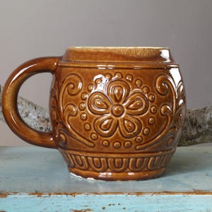 Könnte beinhalten: Eine braune Keramiktasse mit einem Blumendesign. Die Tasse hat einen runden Körper und einen Henkel.