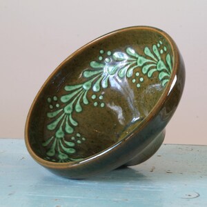 Könnte beinhalten: Eine grüne und braune Keramikschale mit einem stilisierten Blumendesign. Die Schale ist leicht geneigt und hat eine glänzende Oberfläche.